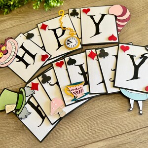 May include: A white and black banner with the word "Happy" spelled out in black letters on white playing card shapes. The banner is decorated with various Alice in Wonderland characters and symbols, including the Cheshire Cat, the Mad Hatter, a teacup, a mushroom, and playing card suits.
