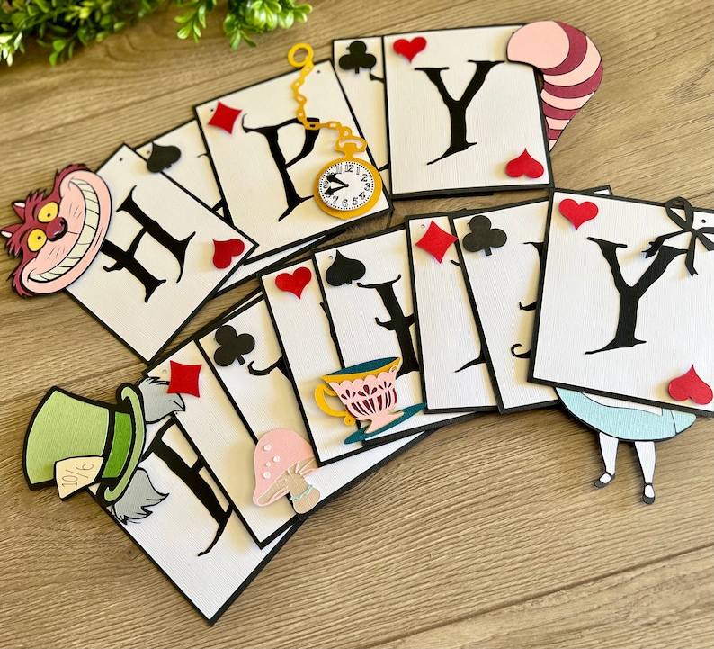 May include: A white and black banner with the word "Happy" spelled out in black letters on white playing card shapes. The banner is decorated with various Alice in Wonderland characters and symbols, including the Cheshire Cat, the Mad Hatter, a teacup, a mushroom, and playing card suits.