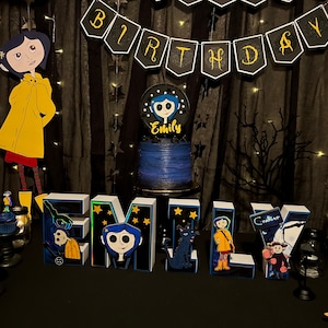 May include: A birthday party table setting with a blue and black cake decorated with a character from the movie "Coraline". The table is decorated with a black and gold "Birthday" banner, black and white stars, and a black and white tree branch. There are also several cupcakes decorated with the same character from the movie.