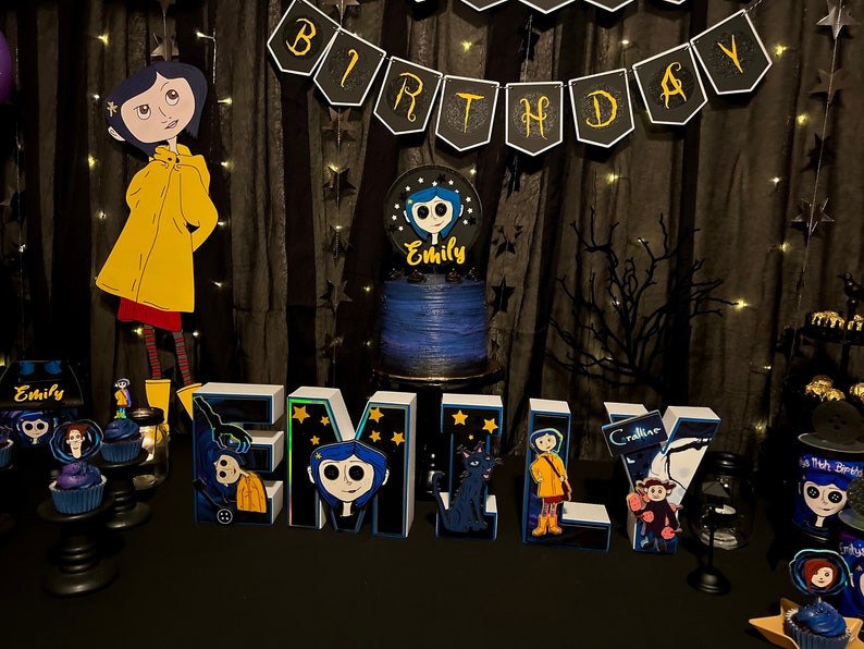 May include: A birthday party table setting with a blue and black cake decorated with a character from the movie "Coraline". The table is decorated with a black and gold "Birthday" banner, black and white stars, and a black and white tree branch. There are also several cupcakes decorated with the same character from the movie.