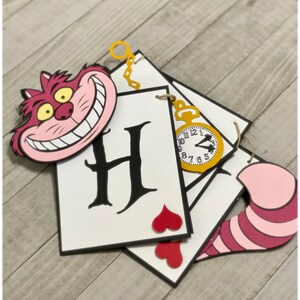 May include: A white card with the letter "H" in black script, a red heart, and a gold pocket watch with Roman numerals. The card is part of a banner with a pink and red Cheshire Cat face and a pink and red tail.