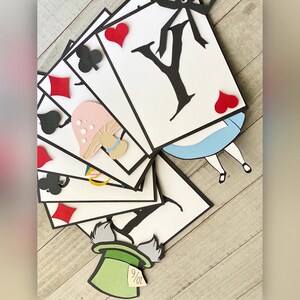 May include: A paper banner with a whimsical Alice in Wonderland theme. The banner features playing cards with black suits and red hearts, a mushroom, a silhouette of Alice, and the Mad Hatter's hat with the number 9/07.