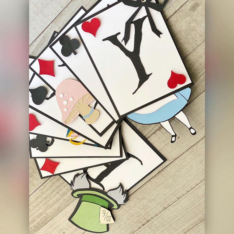May include: A paper banner with a whimsical Alice in Wonderland theme. The banner features playing cards with black suits and red hearts, a mushroom, a silhouette of Alice, and the Mad Hatter's hat with the number 9/07.