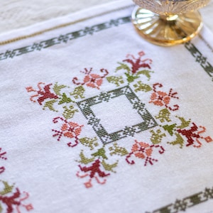May include: White linen placemat with a green and red embroidered floral design. The design features a central square with four corner motifs.