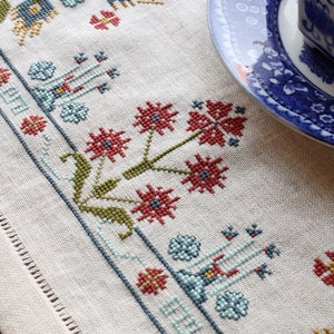 May include: A white linen table runner with a blue, red, and green embroidered floral design. The runner has a fringed edge and is decorated with a repeating pattern of stylized flowers and leaves.