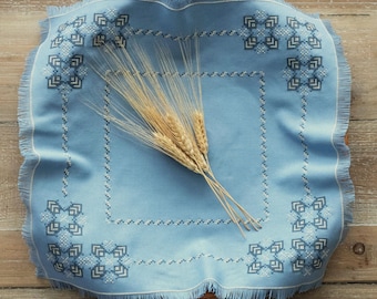 Peut inclure: Une nappe en lin bleu avec une bordure brodée blanche et un motif de petits carrés. Un bouquet d'épis de blé est placé au centre de la nappe.