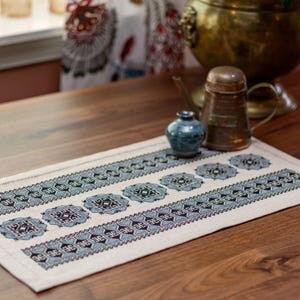May include: A rectangular off-white table runner with a repeating geometric pattern in shades of blue and black. The runner is placed on a wooden table, with a small blue vase and a copper tea pot in the background.