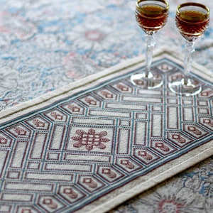 May include: A white table runner with a geometric pattern in shades of blue and red. The runner is made of a woven fabric and has a fringed edge. Two clear glass wine glasses with red wine are on top of the runner.