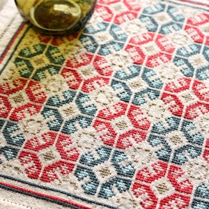 May include: Close-up of a beige embroidered textile with a geometric pattern in red, blue, and green. The design features repeating floral-like shapes. A glass object is partially visible in the upper left corner.
