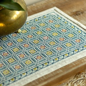 May include: A rectangular, patterned placemat with a geometric design. The pattern features teal, yellow, and red diamond shapes within a grid. The placemat is bordered by a cream-colored edge. A gold-colored decorative bowl is partially visible in the upper left corner.