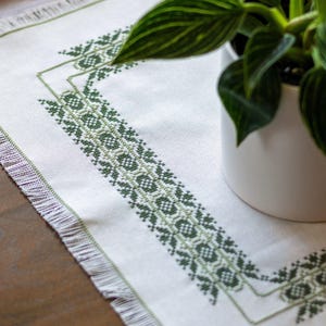 May include: White table runner with a green geometric embroidered design. The runner has a fringed edge and is placed on a wooden surface. A potted plant with green leaves is in the background.