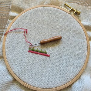 Könnte beinhalten: Ein runder Stickrahmen aus Holz mit einem hellbeigen Stoff. Ein Holz-Punch-Needle-Werkzeug liegt auf dem Stoff, der ein kleines, teilweise fertiggestelltes Stickmuster aus grünen Dreiecken über einer durchgehenden roten Linie zeigt.