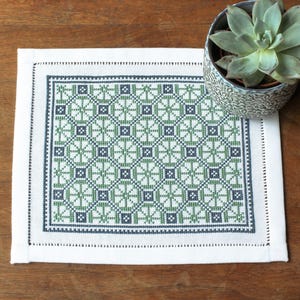 May include: A white linen placemat with a green and blue geometric pattern embroidered in the center. The placemat has a white border with a blue stitched edge.