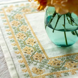 Puede incluir: Un camino de mesa de lino blanco con un borde bordado con motivos florales amarillos y verdes. Un jarrón lleno de rosas amarillas se encuentra sobre el camino de mesa.