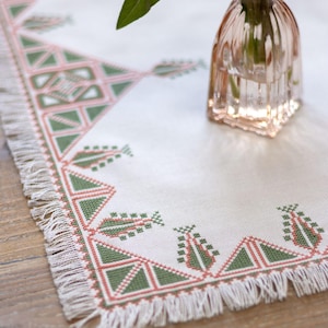 May include: A white linen table runner with a geometric design in green and coral. The runner has a fringed edge and is decorated with a repeating pattern of triangles and other shapes. A small pink glass vase with a green leaf is in the background.
