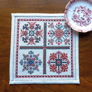 May include: Square, off-white embroidered textile with four geometric floral designs in red, black, and blue. A red and white patterned saucer rests on a wooden surface next to the textile.