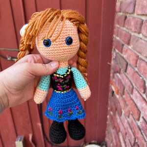 Puede incluir: Mu&ntilde;eca de ganchillo hecha a mano con cabello de hilo naranja peinado en trenzas. La mu&ntilde;eca tiene ojos azules, un tono de piel melocot&oacute;n claro y viste un vestido azul con detalles de flores rosas y un corpi&ntilde;o negro. Los zapatos negros de la mu&ntilde;eca completan el look.