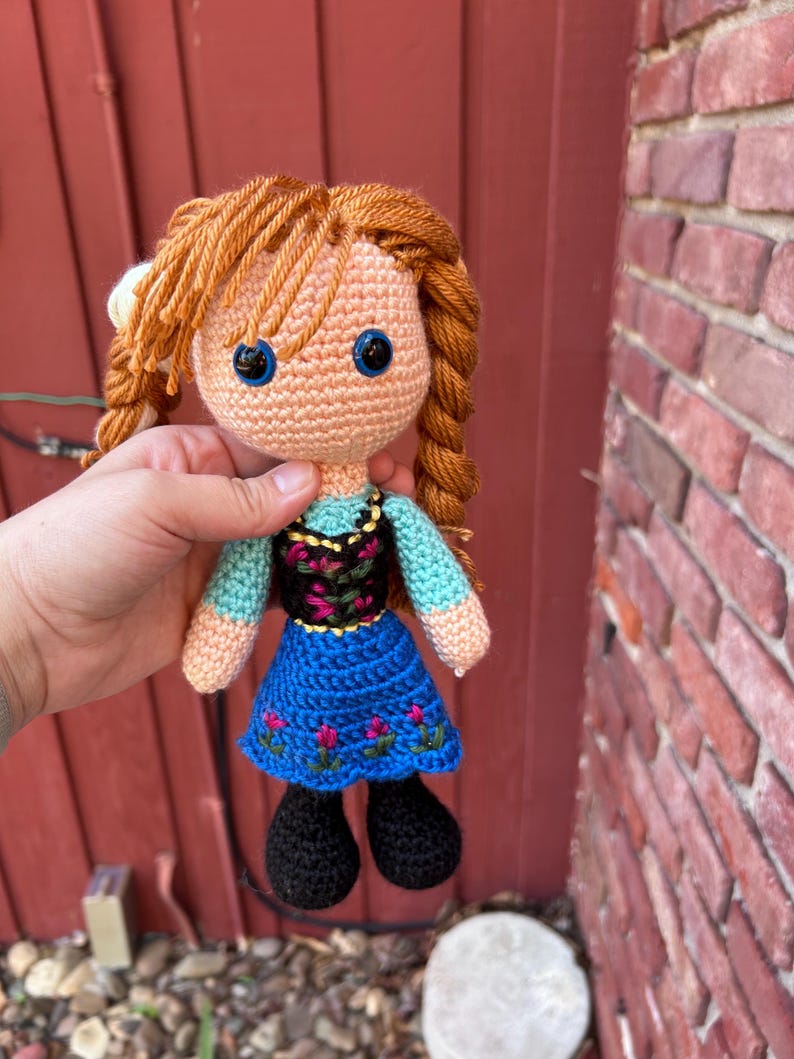 Puede incluir: Mu&ntilde;eca de ganchillo hecha a mano con cabello de hilo naranja peinado en trenzas. La mu&ntilde;eca tiene ojos azules, un tono de piel melocot&oacute;n claro y viste un vestido azul con detalles de flores rosas y un corpi&ntilde;o negro. Los zapatos negros de la mu&ntilde;eca completan el look.