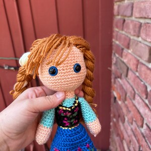 Puede incluir: Mu&ntilde;eca de ganchillo con cabello de hilo naranja peinado en trenzas. La mu&ntilde;eca tiene ojos azules, una blusa azul claro, un corpi&ntilde;o negro con bordado floral y una falda azul con detalles florales. La mu&ntilde;eca lleva zapatos negros.