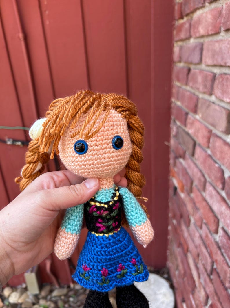 Puede incluir: Mu&ntilde;eca de ganchillo con cabello de hilo naranja peinado en trenzas. La mu&ntilde;eca tiene ojos azules, una blusa azul claro, un corpi&ntilde;o negro con bordado floral y una falda azul con detalles florales. La mu&ntilde;eca lleva zapatos negros.