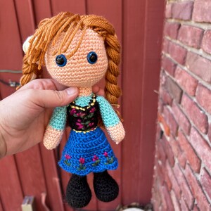 Puede incluir: Mu&ntilde;eca de ganchillo con cabello naranja trenzado. La mu&ntilde;eca tiene ojos azules, una blusa celeste, un corpi&ntilde;o negro con bordado floral y una falda azul con detalles de flores rosas. Zapatos negros completan el atuendo.