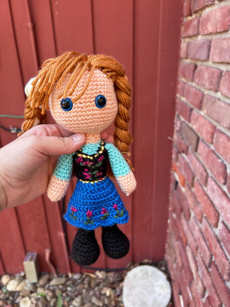 Puede incluir: Mu&ntilde;eca de ganchillo con cabello naranja trenzado. La mu&ntilde;eca tiene ojos azules, una blusa celeste, un corpi&ntilde;o negro con bordado floral y una falda azul con detalles de flores rosas. Zapatos negros completan el atuendo.