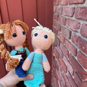 Puede incluir: Dos mu&ntilde;ecas de ganchillo hechas a mano, una con cabello rubio y un vestido azul, la otra con cabello casta&ntilde;o y un vestido azul y negro. Ambas mu&ntilde;ecas tienen ojos azules y est&aacute;n hechas con hilo.