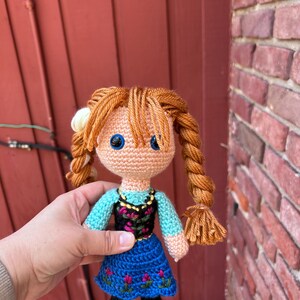 Puede incluir: Mu&ntilde;eca de ganchillo hecha a mano con cabello de hilo naranja peinado en trenzas. La mu&ntilde;eca viste un vestido azul con bordados florales rosas, un corpi&ntilde;o negro y una blusa turquesa. La mu&ntilde;eca tiene ojos azules y zapatos negros.