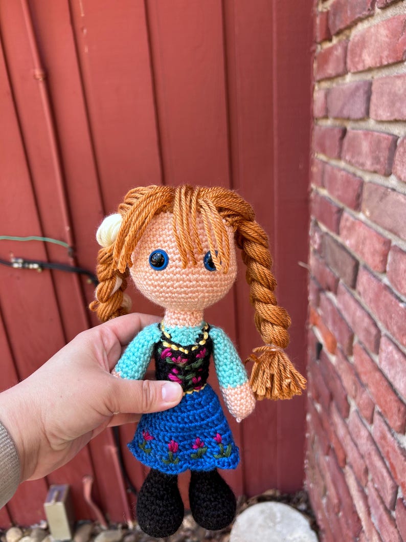 Puede incluir: Mu&ntilde;eca de ganchillo hecha a mano con cabello de hilo naranja peinado en trenzas. La mu&ntilde;eca viste un vestido azul con bordados florales rosas, un corpi&ntilde;o negro y una blusa turquesa. La mu&ntilde;eca tiene ojos azules y zapatos negros.