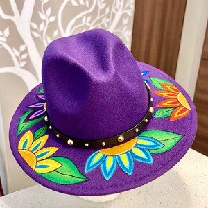 May include: A purple fedora hat with a black band and gold studs. The brim of the hat is decorated with colorful hand-painted flowers.