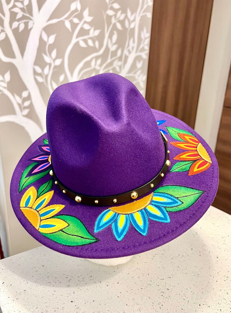 May include: A purple fedora hat with a black band and gold studs. The brim of the hat is decorated with colorful hand-painted flowers.