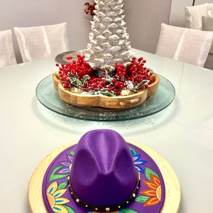 May include: A purple fedora hat with a colorful floral design on the brim, sitting on a wooden base. The hat is decorated with gold studs and a brown leather band.