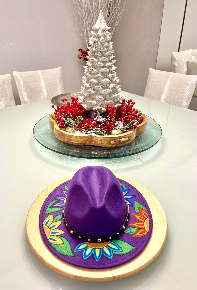 May include: A purple fedora hat with a colorful floral design on the brim, sitting on a wooden base. The hat is decorated with gold studs and a brown leather band.