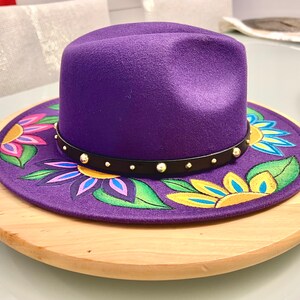 May include: A purple fedora hat with a black band and gold studs. The hat is decorated with hand-painted flowers in shades of yellow, pink, green, and blue.