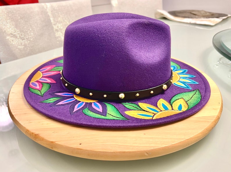 May include: A purple fedora hat with a black band and gold studs. The hat is decorated with hand-painted flowers in shades of yellow, pink, green, and blue.