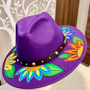 May include: A purple fedora hat with a brown leather band and gold studs. The hat is decorated with colorful hand-painted flowers.