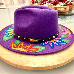 May include: A purple fedora hat with a black band and gold accents. The hat is decorated with colorful flowers painted on the brim.