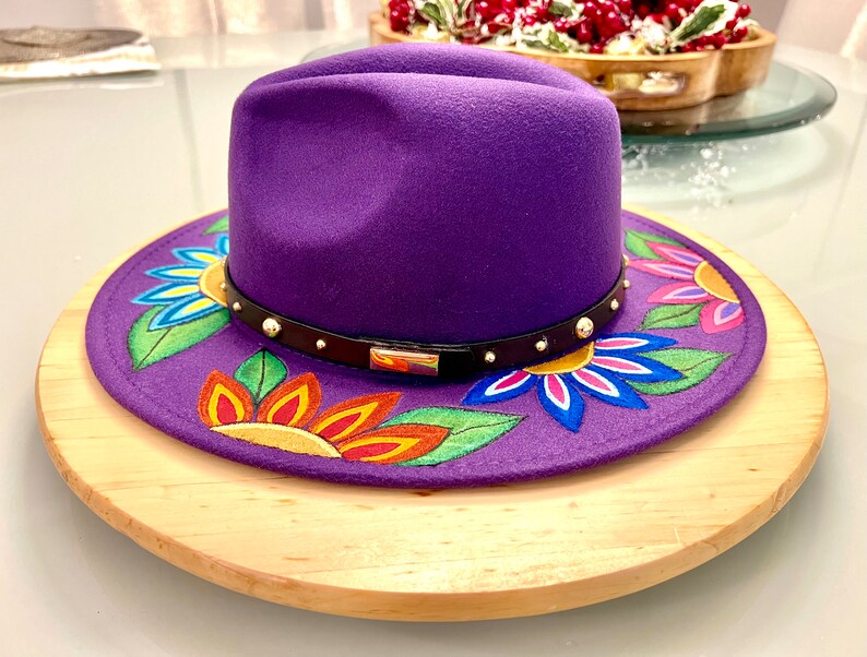 May include: A purple fedora hat with a black band and gold accents. The hat is decorated with colorful flowers painted on the brim.