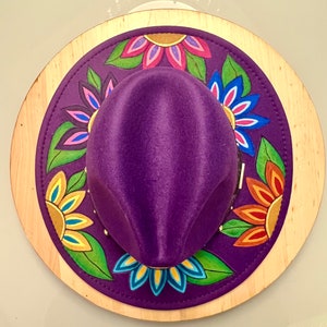 May include: A purple felt hat with a wide brim decorated with colorful painted flowers. The hat is displayed on a wooden base.