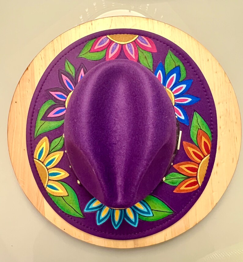 May include: A purple felt hat with a wide brim decorated with colorful painted flowers. The hat is displayed on a wooden base.