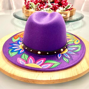 May include: A purple fedora hat with a black band and gold studs. The hat has a colorful floral design painted on the brim.