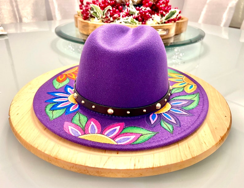 May include: A purple fedora hat with a black band and gold studs. The hat has a colorful floral design painted on the brim.