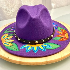 May include: A purple felt fedora hat with a black band and gold studs. The hat is decorated with hand-painted flowers in shades of yellow, orange, red, blue, and green.