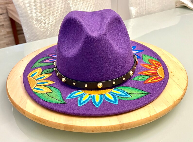 May include: A purple felt fedora hat with a black band and gold studs. The hat is decorated with hand-painted flowers in shades of yellow, orange, red, blue, and green.