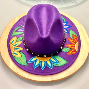 May include: A purple fedora hat with a black band and gold studs. The hat is decorated with painted flowers in shades of yellow, orange, blue, green, and pink.