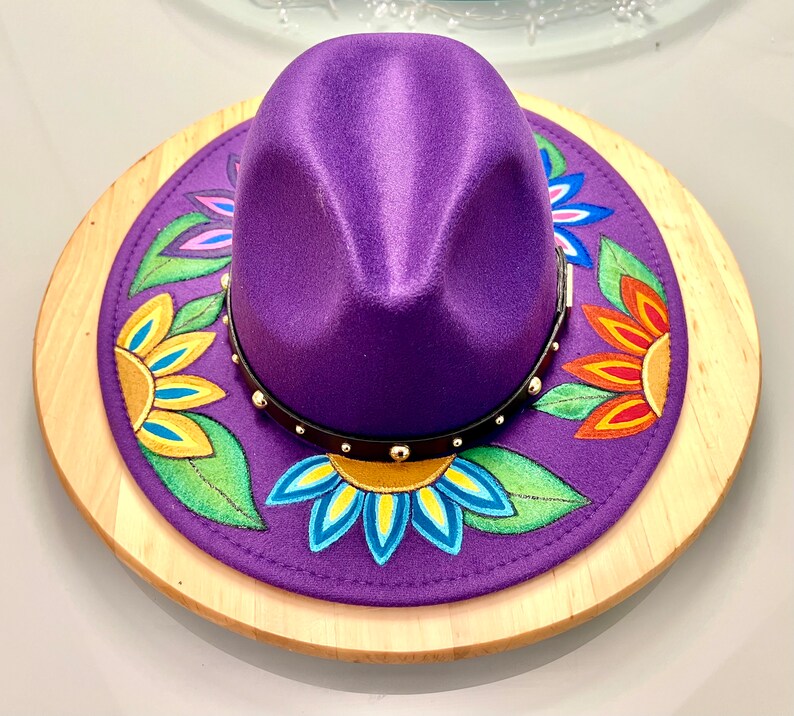 May include: A purple fedora hat with a black band and gold studs. The hat is decorated with painted flowers in shades of yellow, orange, blue, green, and pink.