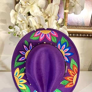 May include: A purple fedora hat with a wide brim decorated with colorful painted flowers. The flowers are yellow, orange, pink, blue, and green.