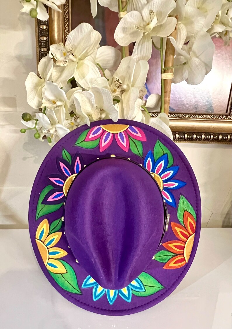 May include: A purple fedora hat with a wide brim decorated with colorful painted flowers. The flowers are yellow, orange, pink, blue, and green.