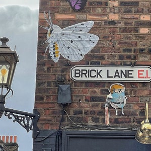 Puede incluir: Un muro de ladrillos con arte callejero, incluyendo una gran polilla con manchas negras y detalles amarillos, y un personaje de dibujos animados. Un letrero de calle dice "BRICK LANE E.1." Una farola ornamentada es visible a la izquierda.