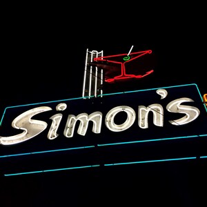 Puede incluir: Letrero de neón para un bar o restaurante llamado "Simon's" con un gráfico de copa de martini sobre el nombre. El letrero está iluminado en blanco, rojo, verde y azul.